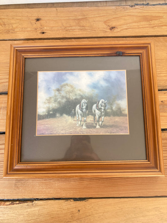A framed art print depicting two horses standing in a field under a cloudy sky, set in a brown wooden frame with a dark mat, displayed on a wooden plank surface.